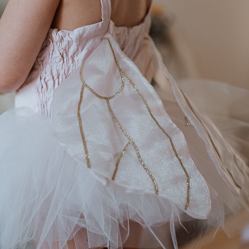 Child wearing a white dress with gold accents and a tutu, standing on a wooden floor.