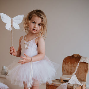 Young girl in a white dress holding a butterfly wand in a room with wooden furniture.