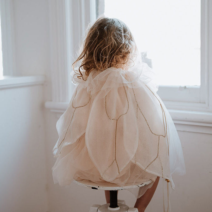 Child wearing a off white fairy costume standing in front of a window.